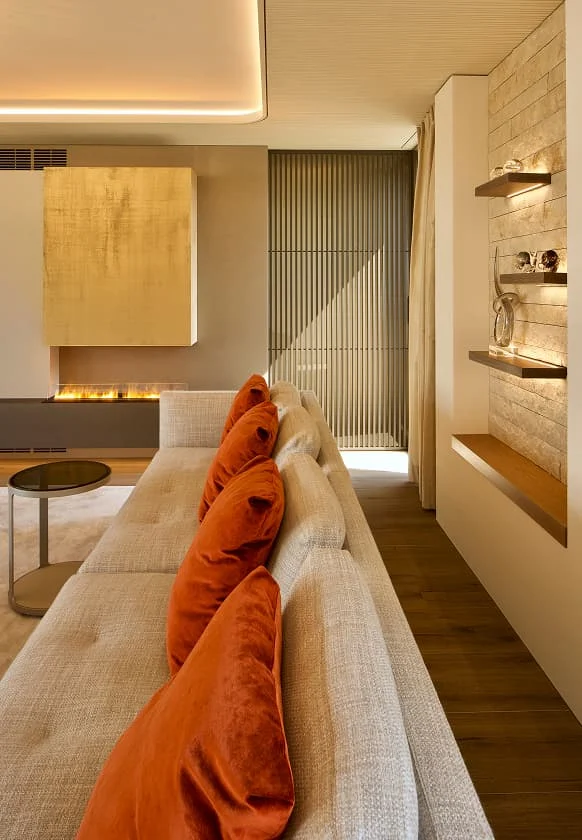 Modernes Wohnzimmer mit cremefarbener Couch, orangefarbenen Samtkissen, offenem Kamin und beleuchteten Wandregalen.