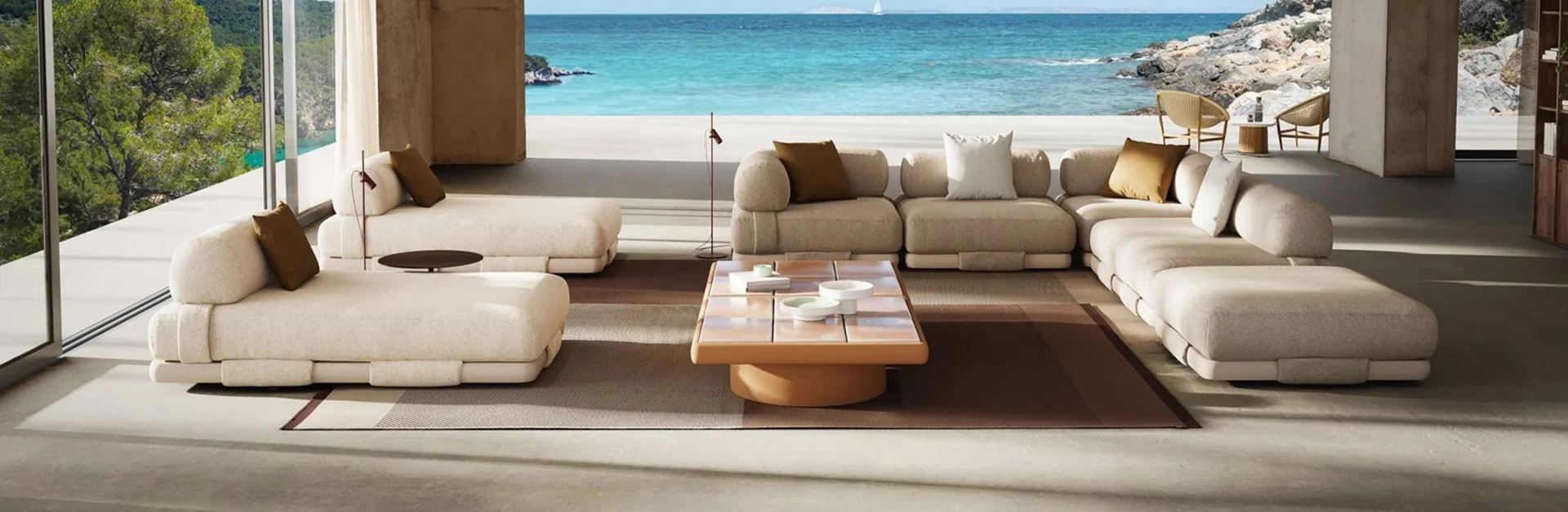 Modernes Wohnzimmer mit großzügigen, beigefarbenen Lounge-Sofas, zentralem Couchtisch und offenem Blick auf das türkisfarbene Meer.