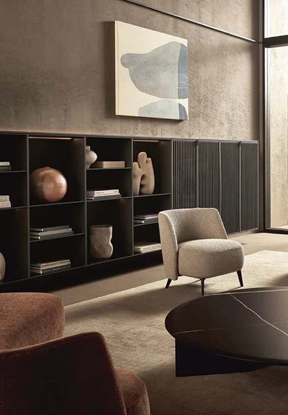 Modernes Wohnzimmer mit offenem Regal, skulpturaler Deko, hell gepolstertem Sessel und rundem schwarzem Couchtisch in warmen Erdtönen.