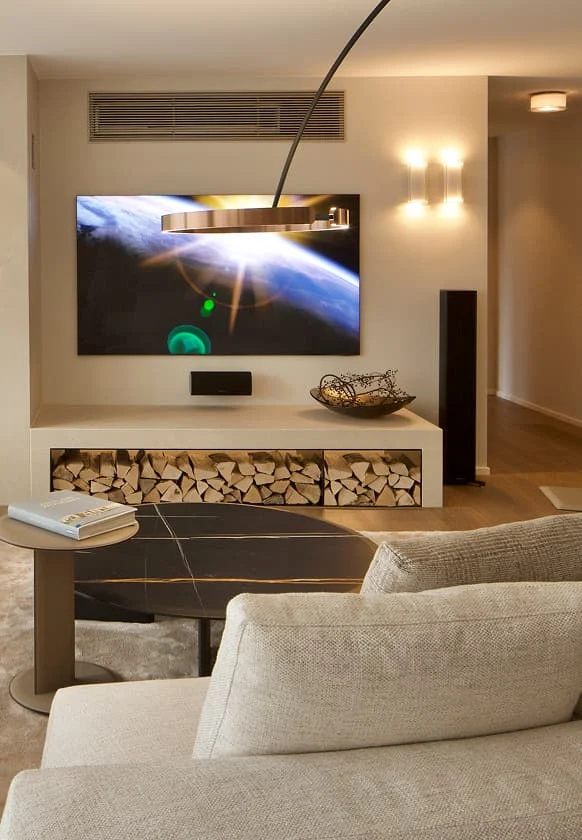 Modernes Wohnzimmer mit großem Wand-TV, dekorativem Kaminholzregal und hellen Polstermöbeln.