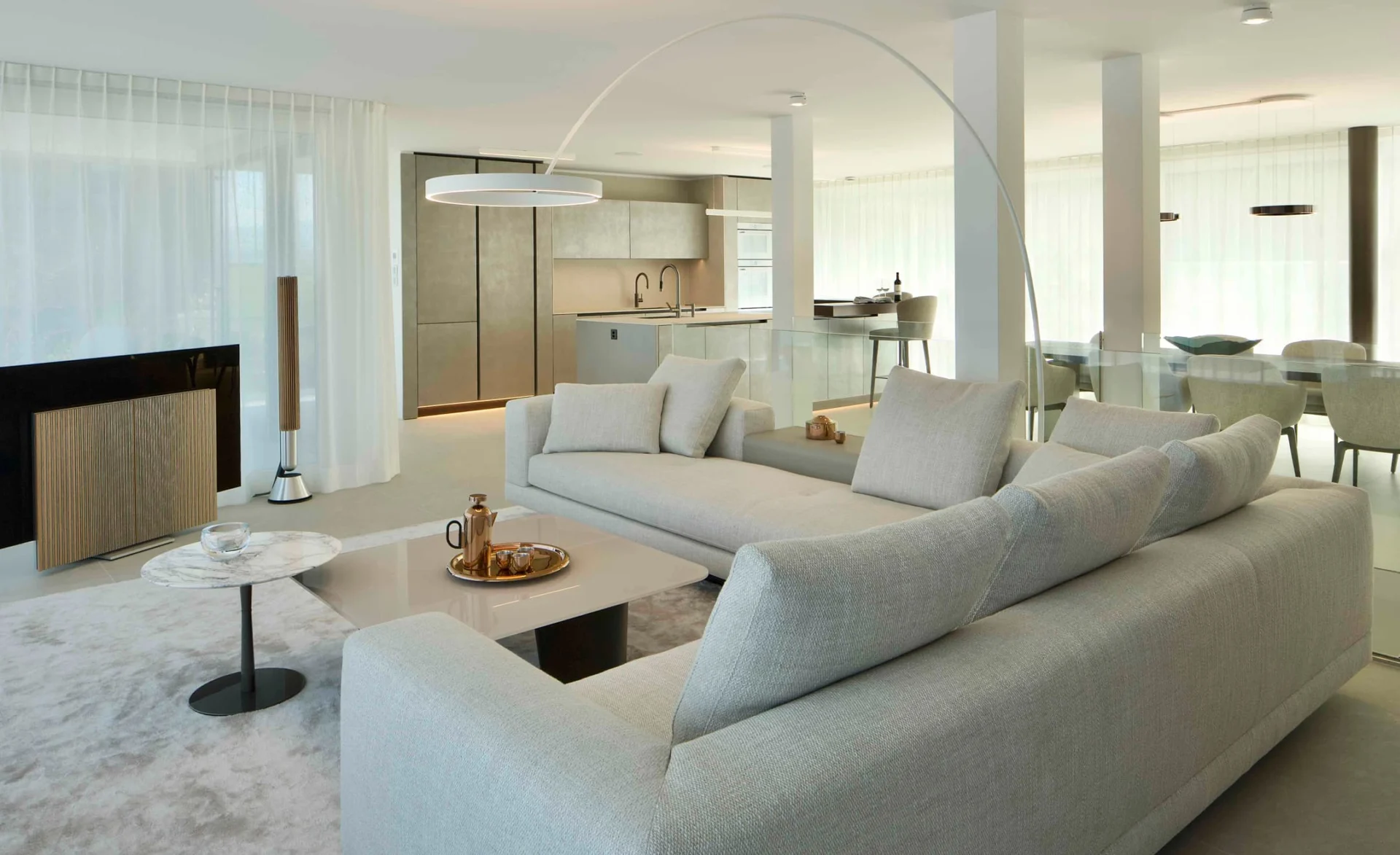 Modernes offenes Wohnzimmer mit großem hellgrauen Sofa, elegantem Esstisch und offener Küche in minimalistischer Einrichtung mit bodentiefen Fenstern.