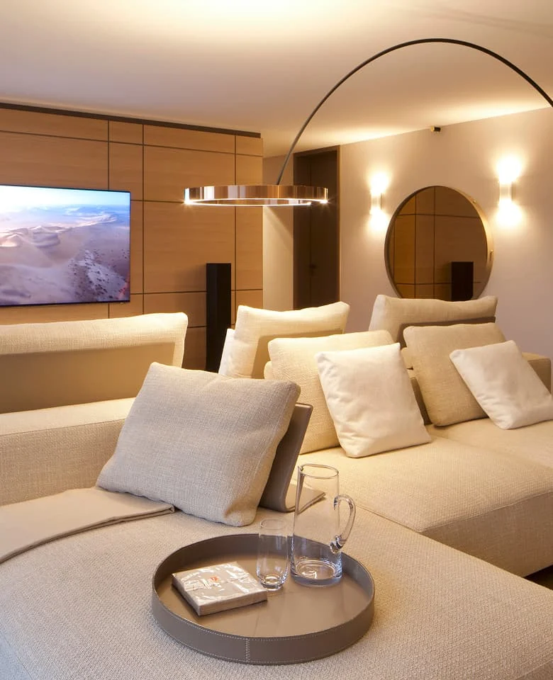 Modernes Wohnzimmer mit großer beiger Sofa-Landschaft, eleganter Ringlampe, Holzpaneelwand mit TV und rundem Spiegel.