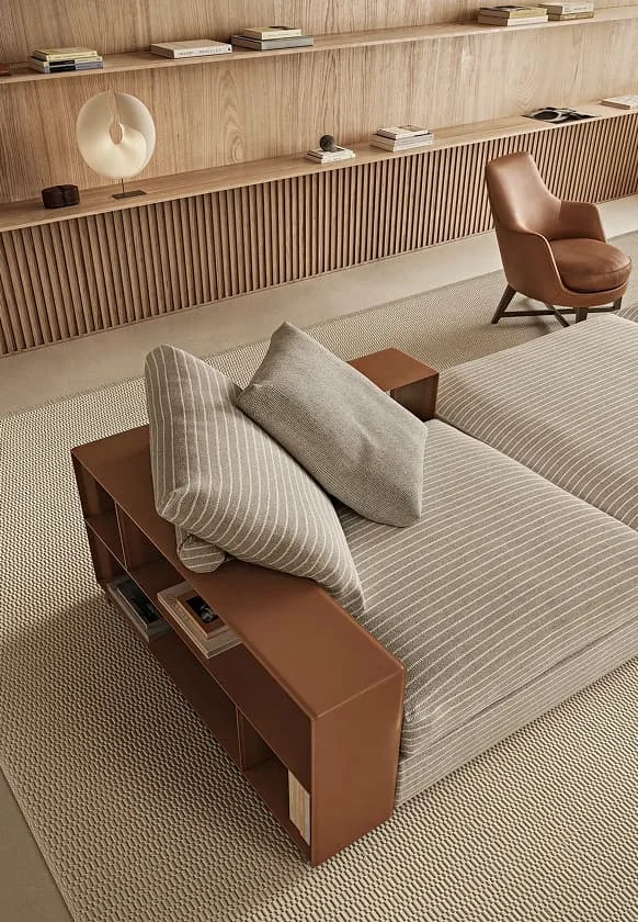 Modernes Wohnzimmer mit modularer Sofalandschaft in Grau, seitlichem braunen Leder-Element mit integrierten Regalfächern, beigem Teppich und Wandregal aus Holz im minimalistischen Design.