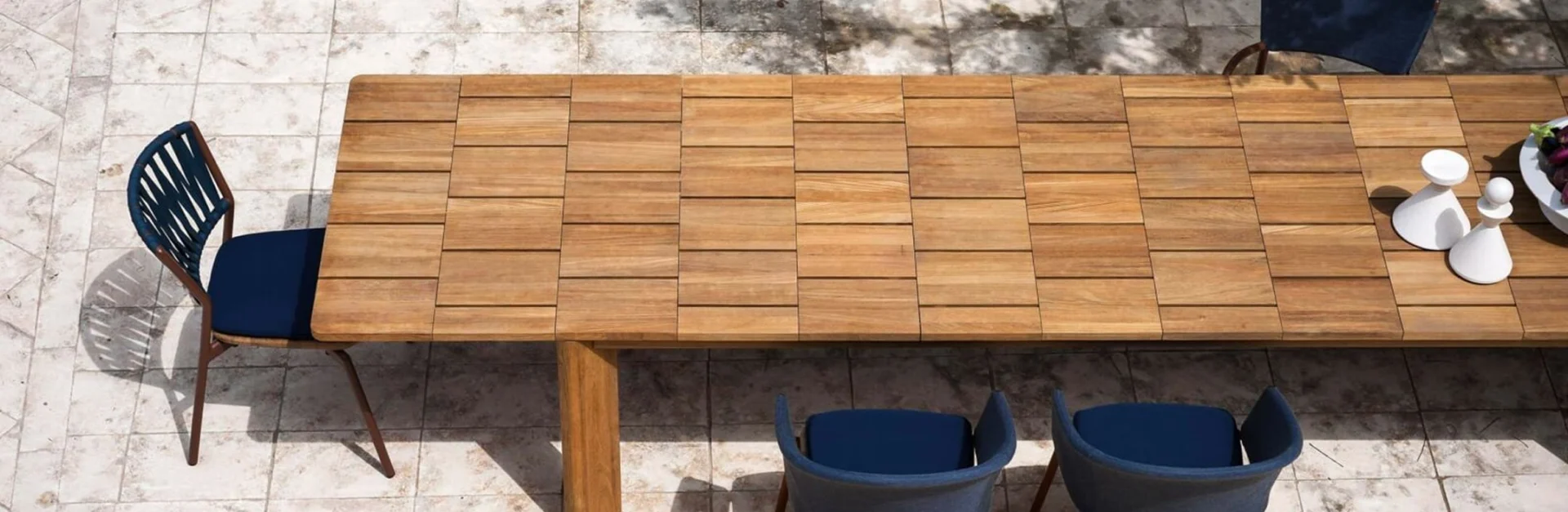 Großer Esstisch aus Holz mit modernem Mosaikmuster auf der Tischplatte, umgeben von blauen Outdoor-Stühlen auf einer hellen Steinterrasse.