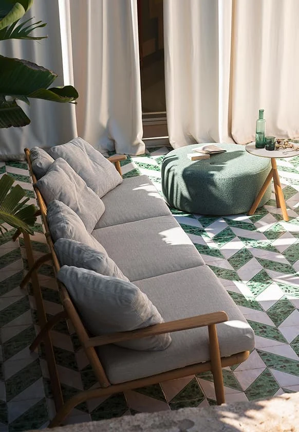 Outdoor-Lounge mit hellgrauem Sofa aus Holzrahmen, grünem Pouf und kleinem Beistelltisch auf gemustertem Terrassenboden in Grün und Weiß.