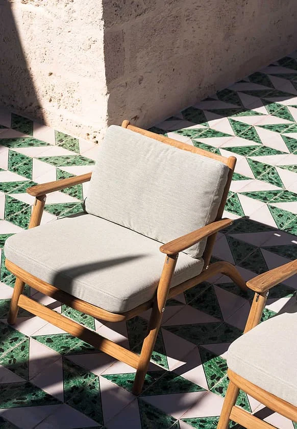 Outdoor-Sessel aus hellem Holz mit beigefarbenen Polstern auf einer Terrasse mit grün-weiß gemusterten Fliesen im Sonnenlicht.