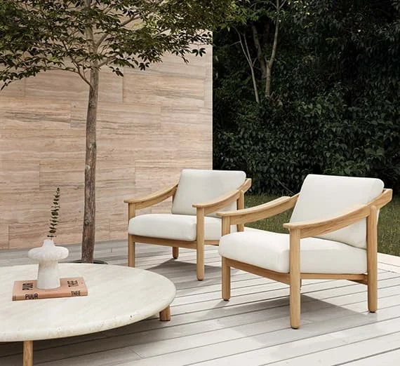 Zwei helle Outdoor-Sessel mit Holzrahmen auf Terrasse neben rundem Steintisch und Baum.