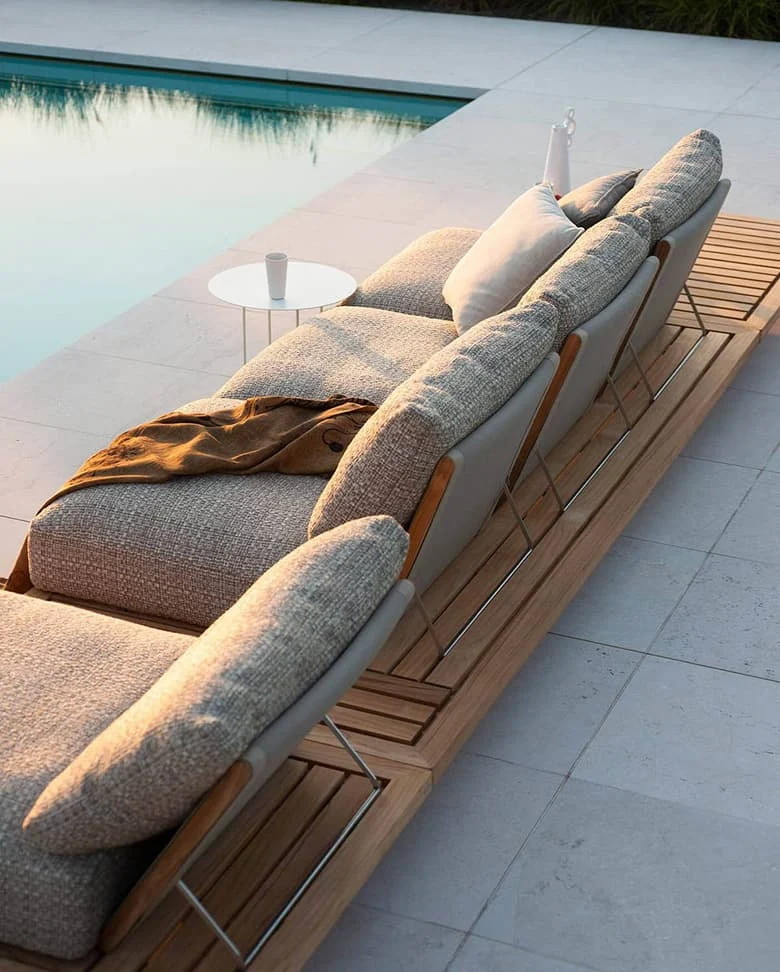 Modernes Outdoor-Sofa aus Holz und Metall mit hellen Polstern neben einem Pool in der Abendsonne.