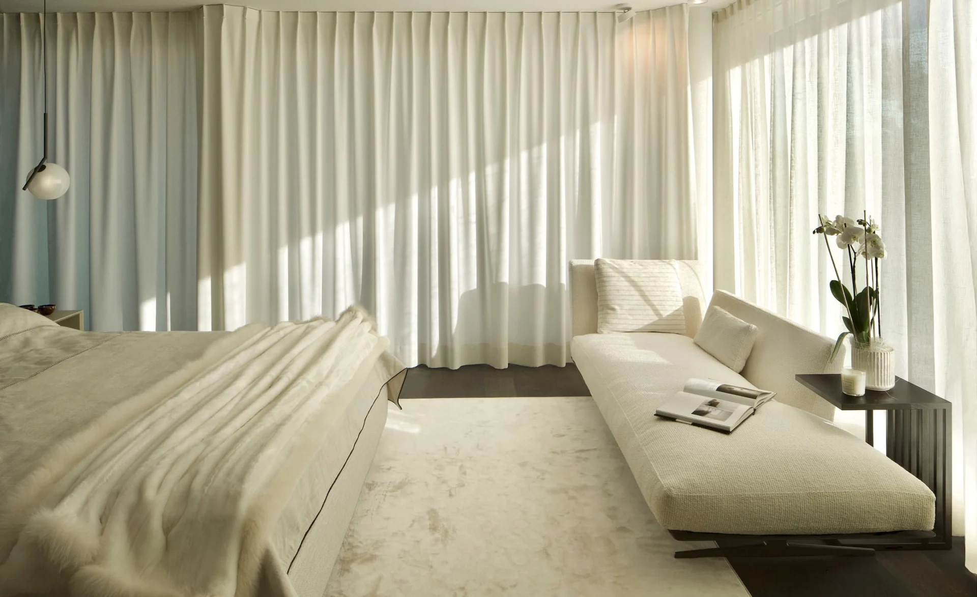 Helles Schlafzimmer mit bodentiefen Vorhängen, cremefarbenem Bett, langer Liege und einer Vase mit weißen Orchideen im Sonnenlicht.