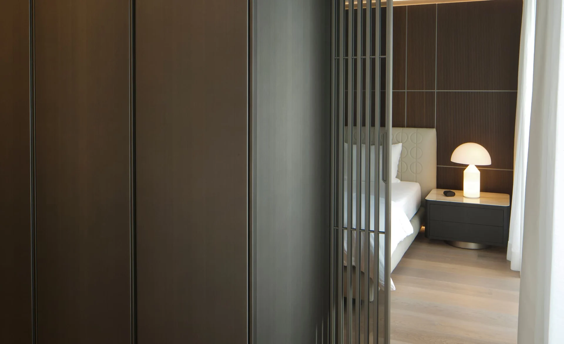 Blick durch eine moderne Lamellen-Schiebetür in ein elegantes Schlafzimmer mit hellem Bett, Holzvertäfelung und beleuchteter Designer-Tischlampe auf dem Nachttisch.