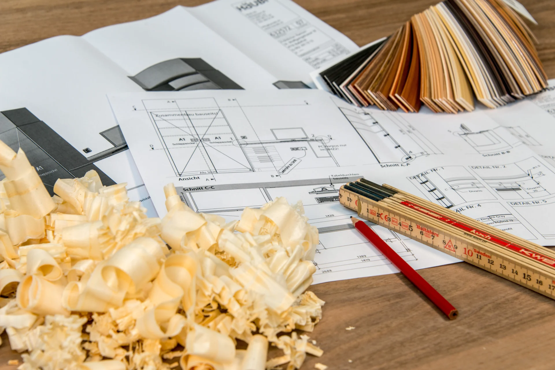 Technische Zeichnungen auf einem Holztisch mit Zollstock, Bleistift, Holzspänen und aufgefächerten Materialmustern.