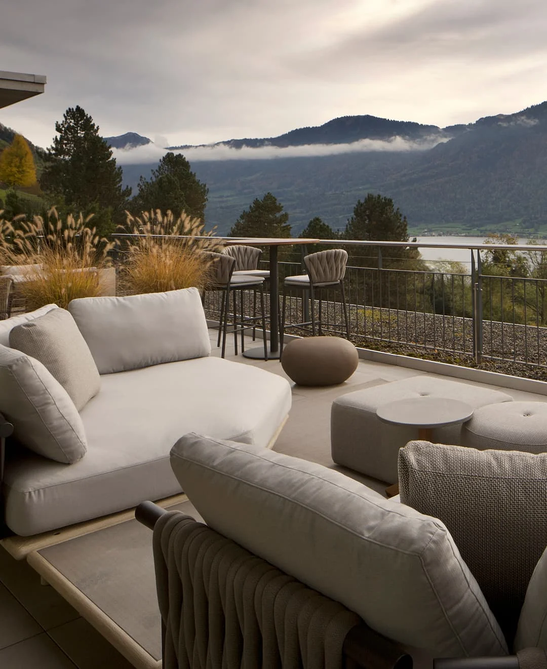 Moderne Terrasse mit gemütlichen Lounge-Möbeln und Blick auf Berge, Wald und einen See im Hintergrund.