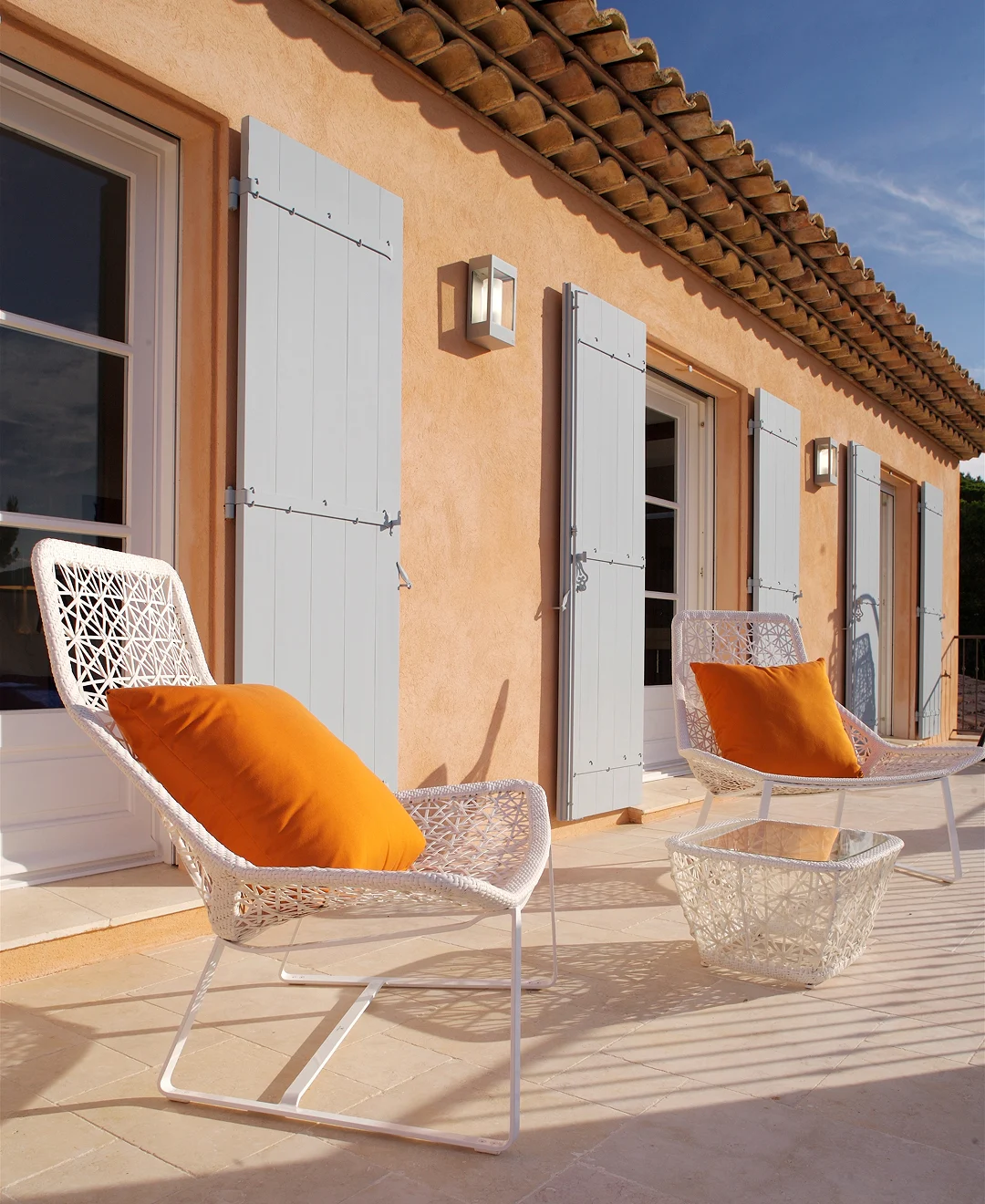 Sonnige Terrasse mit weißen Geflechtstühlen und orangenen Kissen vor einer Terracotta-Hausfassade