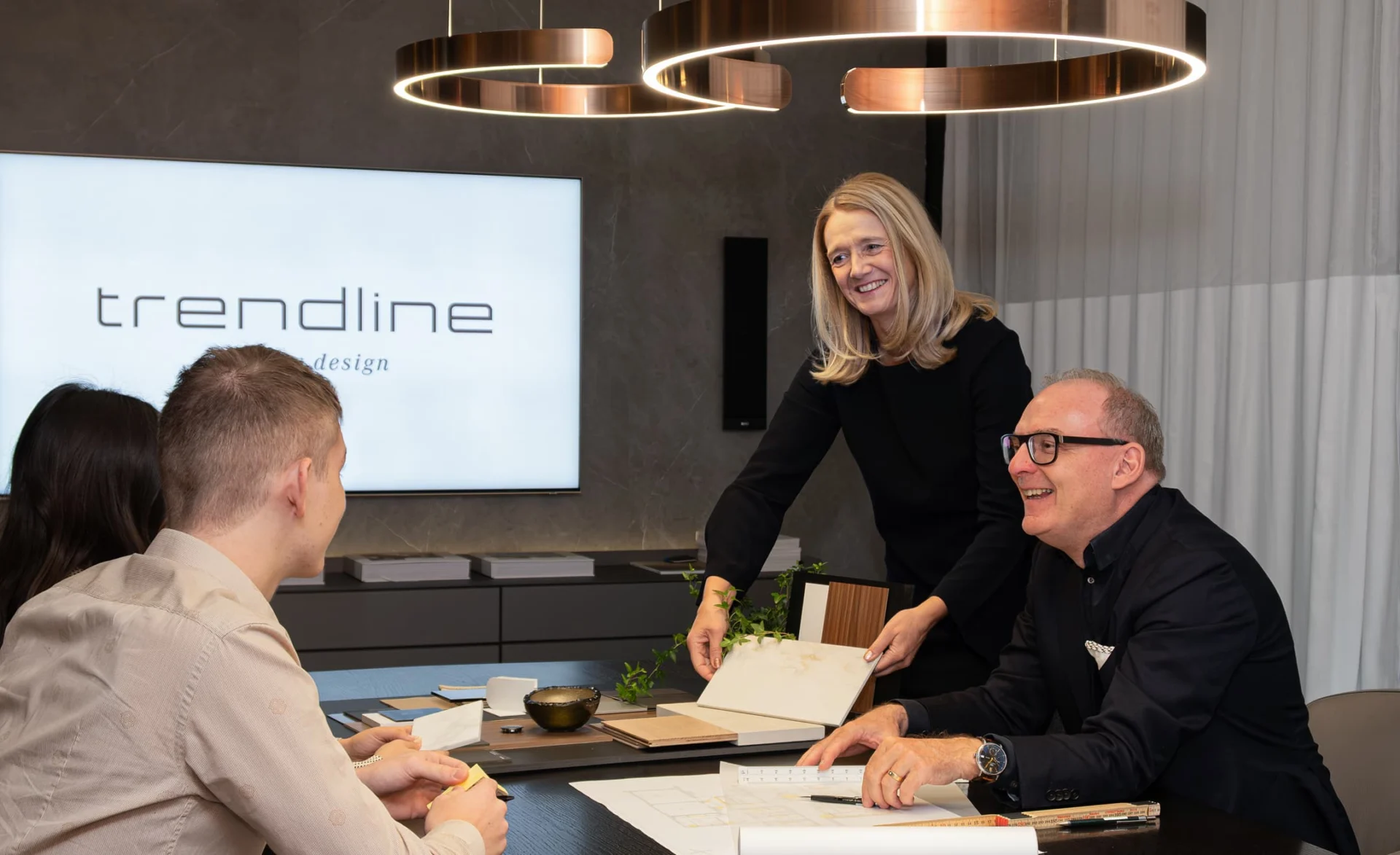 Beratungssituation in einem modernen Innenarchitekturbüro: Eine lächelnde Frau zeigt Materialmuster, während drei Personen am Tisch sitzen und gemeinsam Pläne besprechen. Im Hintergrund leuchtet das Trendline-Logo auf einem Monitor.