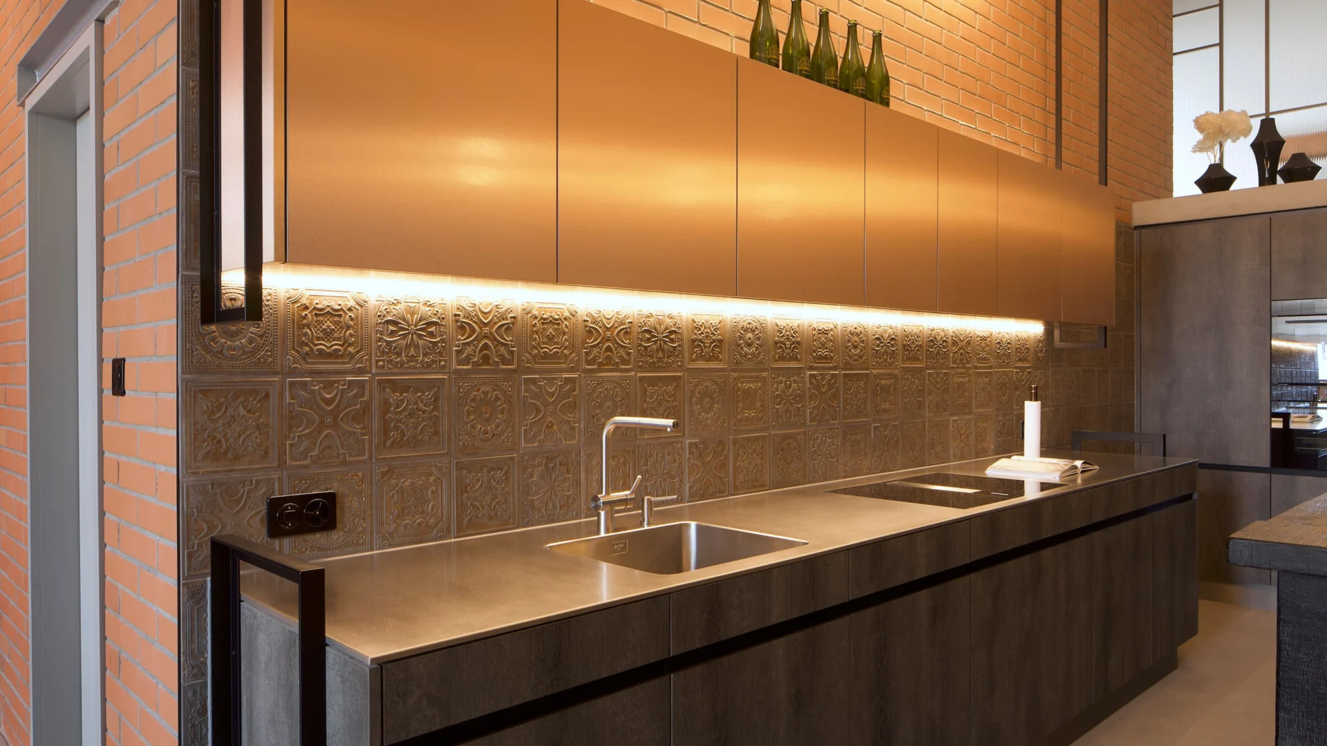 Moderne Küche mit dunklen Fronten, goldener Rückwand und integrierter LED-Beleuchtung – elegantes Zusammenspiel aus warmen Farben, Struktur und industrieller Note.
