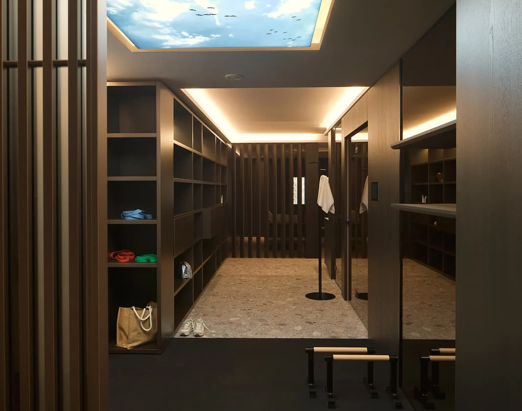 Wellness-Umkleideraum mit großen Holzregalen, indirekter Beleuchtung, heller Steinbodenfläche und einer Lichtdecke mit Himmelmotiv.