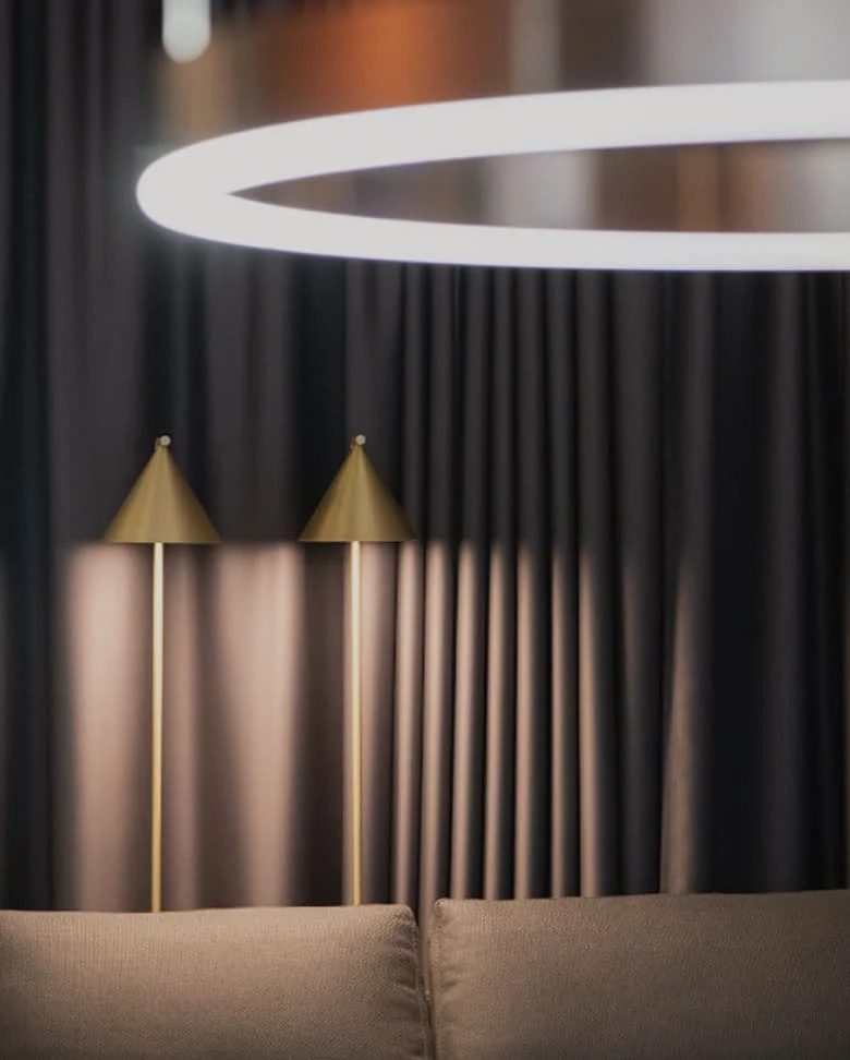 Eleganter Wohnbereich mit modernen Design-Stehlampen in Gold und großer ringförmiger Deckenleuchte – stimmungsvolle Beleuchtung vor dunklem Vorhang.