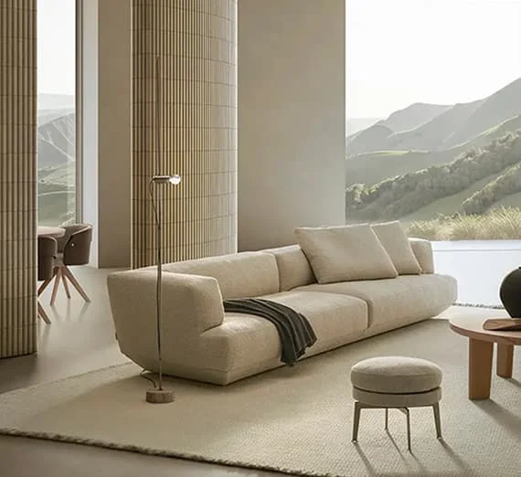 Helles, modernes Wohnzimmer mit beigem Designersofa, rundem Hocker, Stehleuchte und Panoramablick auf grüne Hügel in harmonischer Naturkulisse.