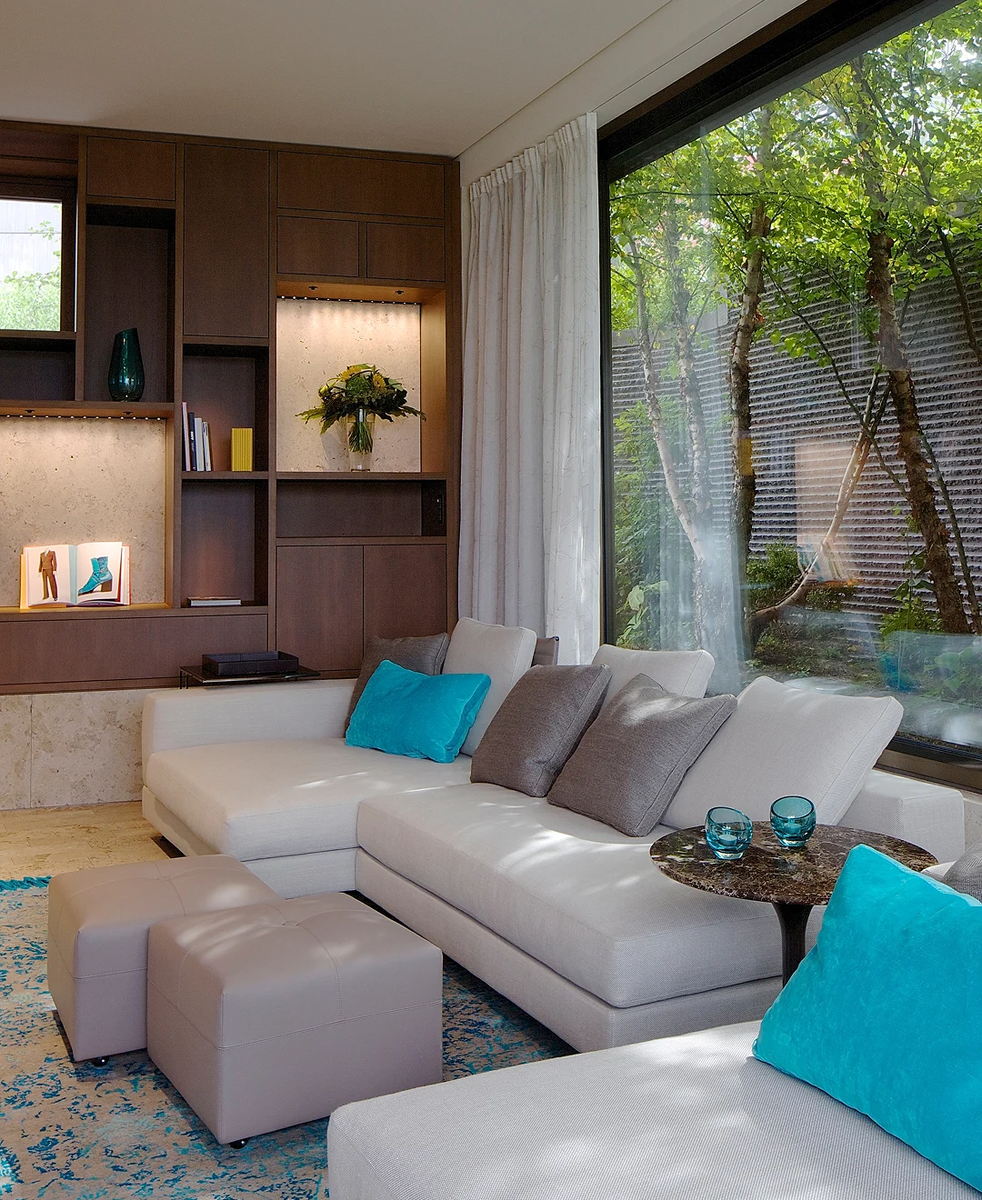 Helles Wohnzimmer mit großem Ecksofa, blauen Kissen, maßgefertigtem Holzregal und Blick ins Grüne durch Panoramafenster.